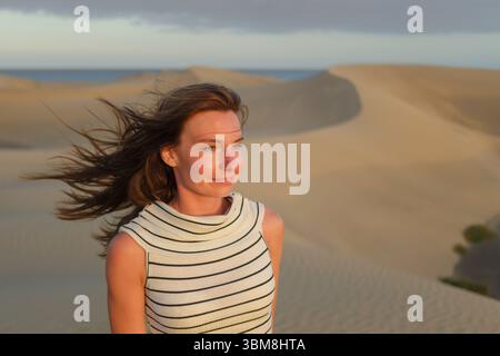 Eine Frau blickt nachdenklich über weite Sanddünen, während die Sonne untergeht, wirft ein warmes Leuchten über die Landschaft und unterstreicht ihre Gesichtszüge. Stockfoto