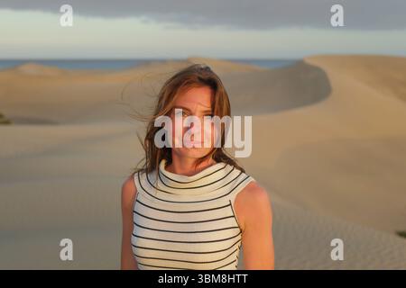 Eine Frau mit langen Haaren posiert in Wüstensanddünen, während die Sonne untergeht, und schafft eine ruhige und malerische Atmosphäre mit einem Strand im Hintergrund. Stockfoto