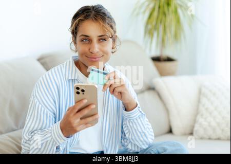 Positive brasilianische Frau, die Kreditkarte und Smartphone hält, Online-Einkäufe tätigt oder Bankgeschäfte von zu Hause aus auf dem Sofa sitzt. Sie sieht beim Einkaufen oder Verwalten von Finanzen auf Mobilgeräten zufrieden und nachdenklich aus Stockfoto