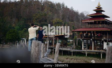 Zwei junge Männer genießen ihren Urlaub und machen Fotos auf einer Holzbrücke mit einem traditionellen Gebäude im Hintergrund Stockfoto