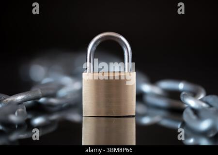 Einfaches Vorhängeschloss aus Metall auf reflektierender schwarzer Oberfläche mit verschwommener Kette im Hintergrund. Nahaufnahme im dunklen Studio mit stimmungsvoller Beleuchtung. Stockfoto