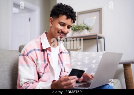 Junger Mann lächelt, während er Laptop und Kreditkarte für Online-Shopping zu Hause benutzt Stockfoto