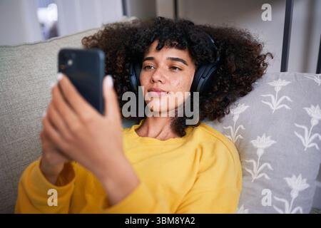 Eine junge Frau, die sich zu Hause mit Kopfhörern und Smartphone entspannt Stockfoto