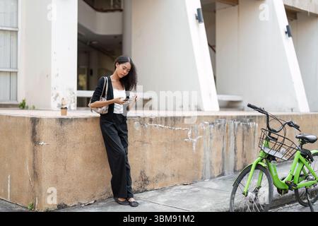 Technologie und Komfort. Eine junge Frau steht lässig, konzentriert auf ihr Smartphone, während sie sich an eine Wand in der Nähe ihres Fahrrads lehnt. Stockfoto