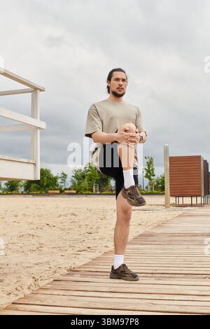 Ein junger Mann mit Bart dehnt sich das Bein auf einem Holzweg am Strand. Stockfoto