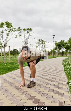 Ein junger Mann mit Bart hockt sich in einem Park und ist bereit, ein Workout im Freien zu beginnen und dabei fit zu bleiben. Stockfoto