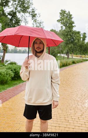Junger Mann in einem Kapuzenpullover, der einen roten Regenschirm hält, während der Regen sanft fällt. Stockfoto