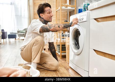 Tätowierter Mann in lässiger Kleidung passt die Waschmaschineneinstellungen zu Hause gerne an. Stockfoto