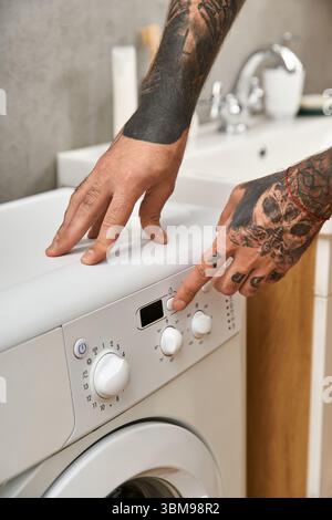 Ein junger Mann mit Tattoos konzentriert sich darauf, die Waschmaschine in seinem komfortablen Zuhause zu stellen. Stockfoto
