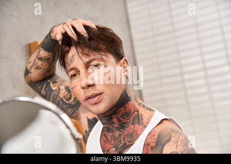 Junger tätowierter Mann in gemütlicher Atmosphäre schaut auf sein Spiegelbild, während er sein Haar anpasst. Stockfoto