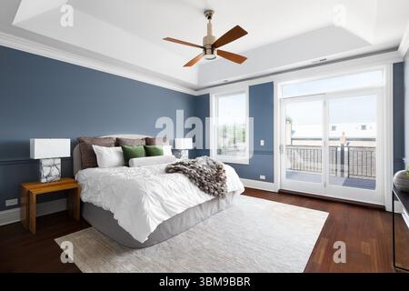 Ein Schlafzimmer mit blauen Wänden, weißer Kassettendecke mit modernem Ventilator, gemütlicher Bettwäsche und Einrichtung sowie dunklem Hartholzfußboden. Stockfoto