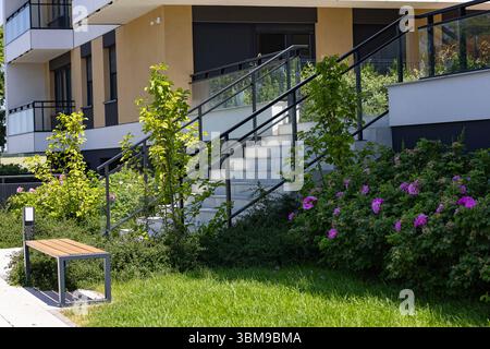 Modernes Wohngebäude mit angelegtem Vorhof, blühenden rosa Blumen, dekorativen Bäumen, Bank und Treppen mit Glasgeländer Stockfoto
