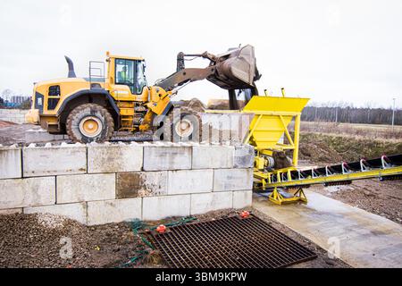 Schwerlast-Frontlader entladen Kies in einen gelben Fördertrichter auf einer Industriebau. Erdbewegungsmaschinen in Aktion auf einem Werk Stockfoto