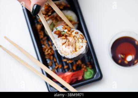 Sushi-Rolle, die von Stäbchen gehalten wird. Asiatisches Essen mit Reis und Gemüse. Sojasauce und Holztisch im Hintergrund. Schwarzes Tablett mit Sushi zum Mitnehmen. Stockfoto