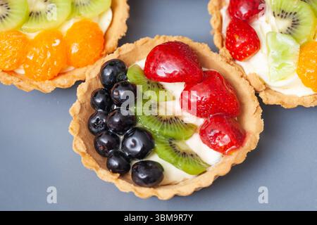 Bunte Obstkuchen auf blauem Hintergrund. Mini-Dessertgebäck mit Kiwi-Erdbeere, Heidelbeere und Orange. Frischer süßer Snack mit Vanillepudding und Glasur. Stockfoto