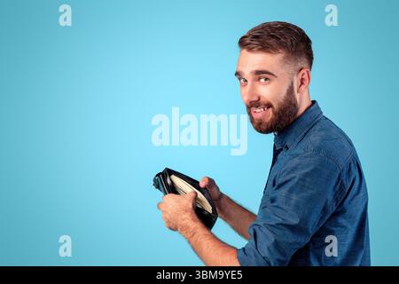 Funky kaukasischen Kerl hält volle Brieftasche mit Geld auf blau studio-Hintergrund Stockfoto