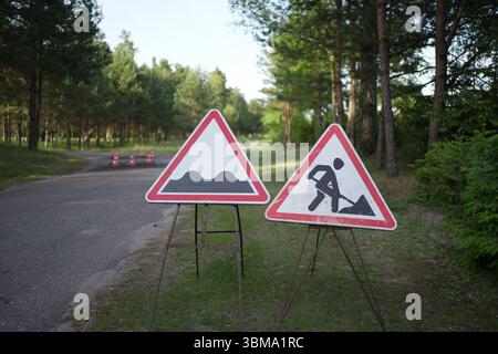 Zwei rote dreieckige Straßenschilder auf einer bewaldeten Landstraße, die auf eine holprige Straße hinweisen und vor Bauarbeiten warnen. Stockfoto