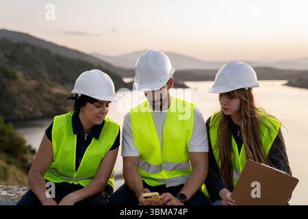 Ingenieure mit Smartphone in der Nähe des Sees bei Sonnenuntergang: Teamarbeit und Technologie in Infrastrukturprojekten Stockfoto