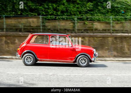 Klassischer roter Mini Cooper, der auf einer Straße mit einer Steinmauer und Grün im Hintergrund fährt Stockfoto