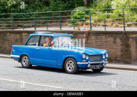 Ein klassischer blauer Triumph Herald aus den 1960er Jahren, der auf einer Stadtstraße mit einer Backsteinmauer und Grün im Hintergrund fährt Stockfoto