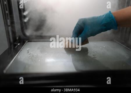Professionelle Reinigungskraft trägt blauen Handschuh mit Schwamm zum Reinigen der Glastür des Gasofens in dunkler Küche, Nahaufnahme zugeschnittener Schuss. Stockfoto
