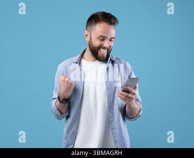 Porträt von glücklichen bärtigen jungen Mann mit Smartphone, so dass Gewinner Geste über blauen Studio-Hintergrund Stockfoto