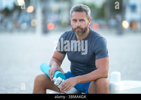 Sport-Fitness-Mann, der draußen auf einer Bank ruht. Reifes Fitnesssportmodell, das nach dem Training auf einer Bank sitzt. Senior Sports man macht danach eine Pause Stockfoto
