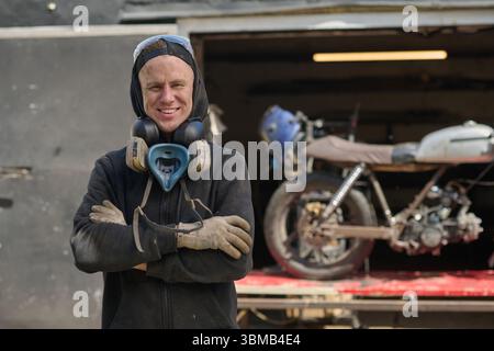 Kaukasischer junger Erwachsener lächelt beim Zusammenbauen eines Custom Motorrads in der Werkstatt Stockfoto