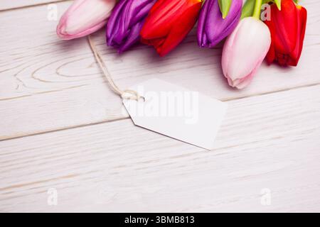 Das flache Design zeigt Tulpenblüten auf einer hellen Holzplatte mit leerem weißem Geschenkanhänger Stockfoto