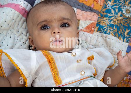 Niedliches indisches Baby in traditionellem weißem Kleid mit gelbem Besatz, das auf farbenfroher Decke liegt und neugierig in die Kamera blickt Stockfoto