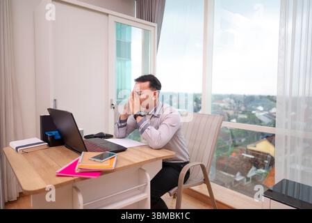 Indonesischer südostasiatischer Geschäftsmann, der Stress und Burnout erlebt, während er an einem hybriden Arbeitsplatz an einem Laptop arbeitet. Papierkram und ein Handy auf dem T Stockfoto