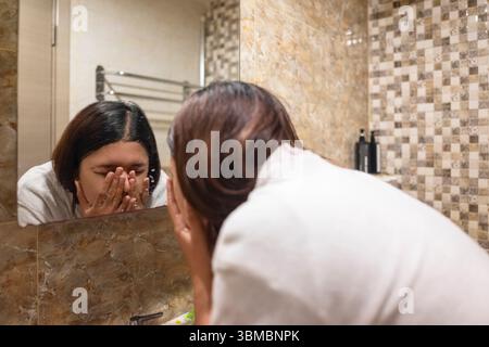 Eine indonesische südostasiatische Frau, die in ein Handtuch gewickelt ist und ihr Gesicht mit Spritzwasser wäscht und reinigt. Natürliche Hautpflege und Wellness-Konzept Stockfoto
