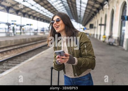 Junge Frau, die ihr Smartphone benutzt, während sie am Bahnhof auf einen Zug wartet, lächelt und ihr Reiseabenteuer genießt Stockfoto