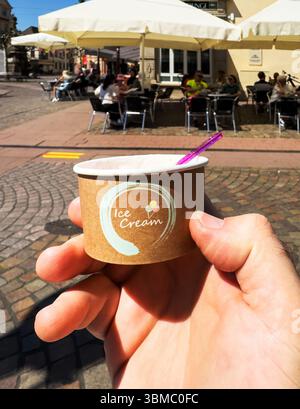 Rastatt, Deutschland - 1. Mai 2025: Eine Hand hält einen kleinen Eisbecher mit einem pinkfarbenen Löffel im sonnigen Zentrum von Rastatt, Deutschland, mit Menschen, die sich entspannen Stockfoto
