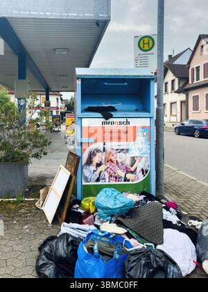 Ulm, Deutschland - 6. Mai 2025: Ein Textilrecyclingbehälter an einer Bushaltestelle in einer deutschen Stadt wird von weggeworfener Kleidung und Taschen umgeben, mit Spende Stockfoto