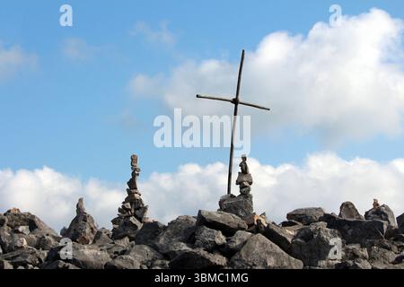Ein einfaches Holzkreuz steht zwischen ausgeglichenen felsenhügeln auf einem felsigen Hügel vor einem hellblauen Himmel mit verstreuten Wolken, die den Glauben symbolisieren Stockfoto