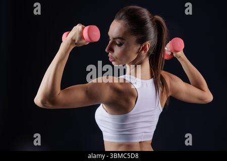 Sportliche Frau in Sportbekleidung beugt Rücken- und Armmuskeln mit Kurzhanteln, zeigt Kraft und Fitness im Studio Stockfoto