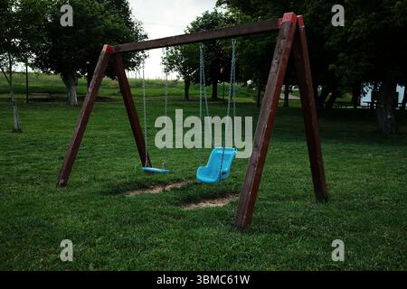 Zwei leere Schaukeln, darunter ein Kindersitz, hängen an einem Holzrahmen in einem ruhigen grünen Park. Kleine serbische Stadt Stockfoto