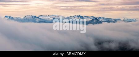 Schneebedeckte Gipfel der Berge steigen bei Sonnenaufgang aus dicken Wolken hervor und bieten einen atemberaubenden Blick. Das sanfte Morgenlicht verstärkt die ruhige Atmosphäre Stockfoto