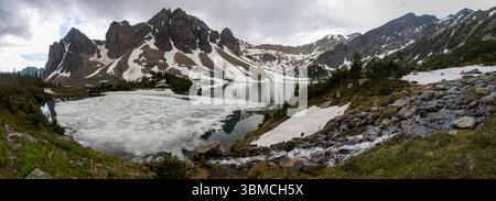 Eine malerische alpine Szene bietet einen ruhigen See, der von schneebedeckten Bergen umgeben ist. Geschmolzener Schnee bedeckt Teile der Landschaft, mit grünem Auftauchen Stockfoto