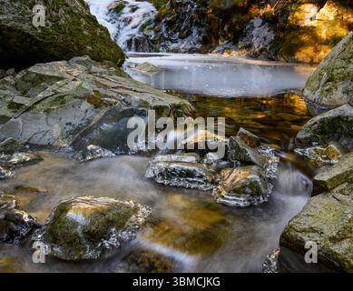 Ein ruhiger Fluss fließt sanft über glatte Felsen, umgeben von üppigem Grün und beleuchtet von sanftem Morgenlicht. Das Wasser glitzert, wenn es kaskadiert, c Stockfoto