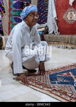 Marokko, Merzouga, 4. Juli 2025. Berber man präsentiert einen traditionellen handgefertigten Teppich in einem marokkanischen Teppichgeschäft, in dem das reiche Kunsthandwerk der Region präsentiert wird Stockfoto