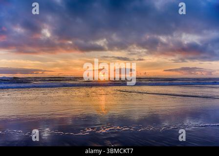 Wellen schlagen sanft gegen die Küste, während die Sonne am Horizont untergeht, warme Farben über den Himmel wirft und sich auf dem nassen Sand des Strandes reflektiert. Stockfoto