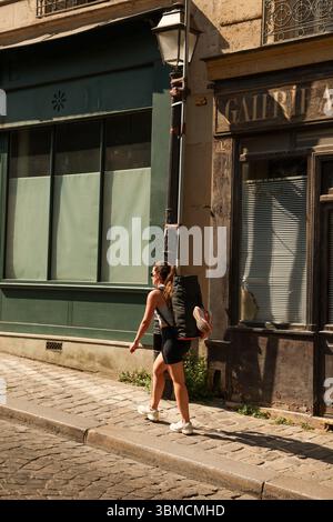Eine junge Frau mit Sonnenbrille läuft eine sonnige Kopfsteinpflasterstraße in Paris hinunter, mit einer großen schwarzen Reisetasche. Die Szene wird von einem leicht aufgenommen Stockfoto