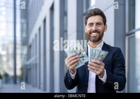 Ein erfolgreicher Geschäftsmann, der Bargeld hält und selbstbewusst vor einem Bürogebäude lächelt, ein Symbol für Reichtum. Stockfoto