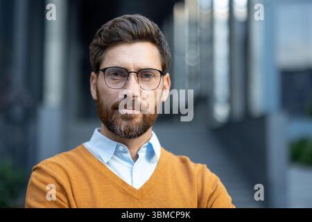 Ein Porträt eines Mannes mit Brille und Bart, der einen gelben Pullover und ein hellblaues Hemd trägt, im Freien aufgenommen. Stockfoto