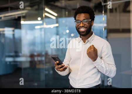 Ein aufgeregter Mann lächelt, während er ein Smartphone hält, und feiert einen Erfolg in einem modernen Büroumfeld. Stockfoto