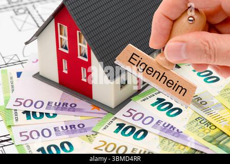 Eigenheim und Eigentum mit Finanzierung ein Holzstempel in der Hand ist beschriftet mit dem Aufdruck Eigenheim und wird vor einem Modellhaus mit vielen Stockfoto