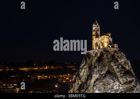 Nächtlicher Blick auf die Kapelle von Saint-Michel d'Aiguilhe auf einem felsigen Hügel, die von den Lichtern der Stadt umgeben ist, die unter dem dunklen Himmel in Aiguilhe, Frankreich, leuchten. Stockfoto
