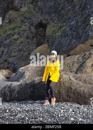 Eine Frau in einer gelben Jacke hebt sich von den dunklen Vulkanfelsen am Strand von Reynisfjara ab. Die Sonne wirft Schatten, während die Meereswellen sanft abstürzen und die einzigartige vulkanische Landschaft Islands hervorheben. Stockfoto